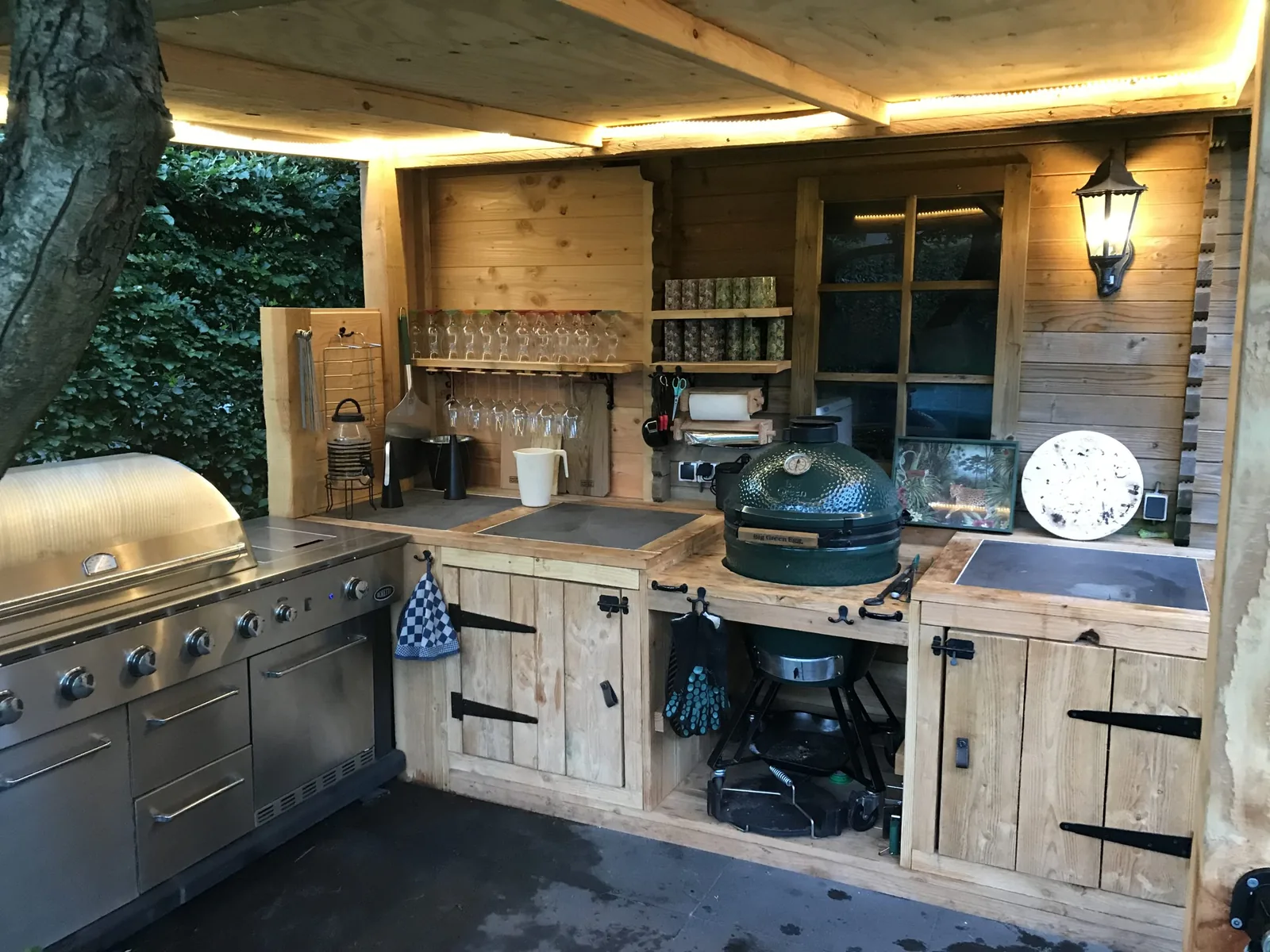 Zelfgebouwde buitenkeuken van Eduard Renger: houten maatwerk-kasten met stalen grepen, een gasgrill en een Big Green Egg onder een houten overkapping